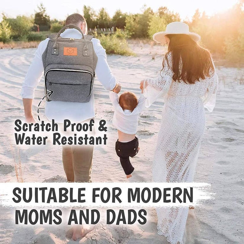 May include: A gray diaper bag backpack with a small bear logo and the text "Scratch Proof & Water Resistant". The bag is being worn by a person walking on a sandy beach with a child and another person.