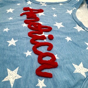 May include: A light blue sleeveless shirt featuring a white star pattern throughout the fabric. The word America is written vertically in a textured, raised red script font across the center of the garment, creating a bold contrast.