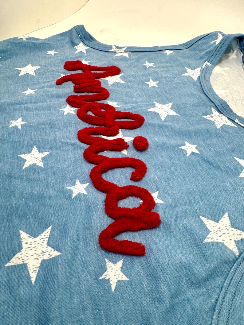 May include: A light blue sleeveless shirt featuring a white star pattern throughout the fabric. The word America is written vertically in a textured, raised red script font across the center of the garment, creating a bold contrast.