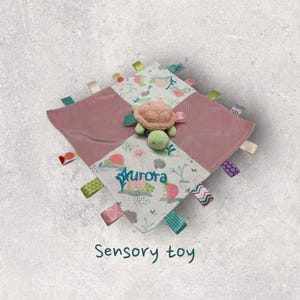 May include: A pink and white sensory toy with a green and pink turtle in the center. The toy has a variety of textures and colors, including pink, green, white, and red. The toy is personalized with the name "Aurora".