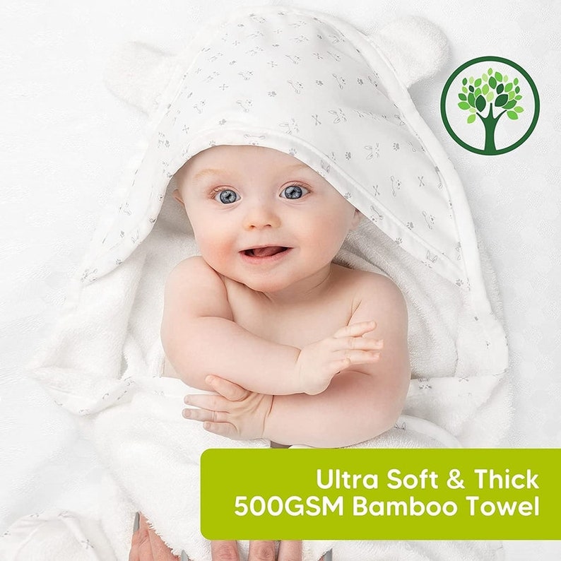 May include: A baby wrapped in a white hooded towel with a pattern of small gray shapes. The towel has bear-like ears on the hood. The text "Ultra Soft & Thick 500GSM Bamboo Towel" is in a green rectangle.