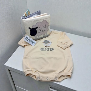 May include: A light beige baby romper with a sheep graphic and the text "CHILD OF GOD". A small fabric book with the text "Precious Little One, Always Remember" and "You are a Child of God" is next to the romper. A tag with the name "Rhenn" is attached.