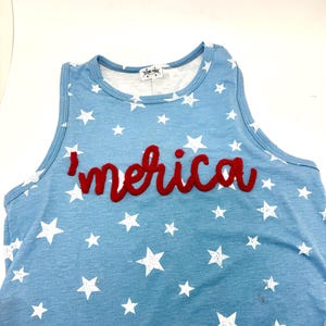 May include: A light blue sleeveless tank top featuring a white star pattern throughout. The word 'merica' is embroidered in red cursive script across the chest. A white label with the text 'TEXAS-TRUE' is visible at the inside neckline.
