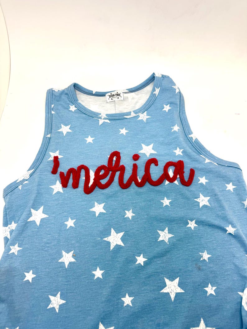 May include: A light blue sleeveless tank top featuring a white star pattern throughout. The word 'merica' is embroidered in red cursive script across the chest. A white label with the text 'TEXAS-TRUE' is visible at the inside neckline.