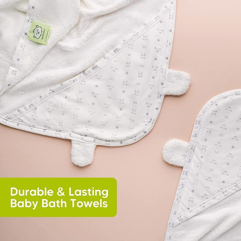 May include: White baby bath towels with a hood and a pattern of small gray bunny faces and paw prints. The towels have a soft, plush texture and a tag with a green logo. The text "Durable & Lasting Baby Bath Towels" is displayed in a green rectangle.