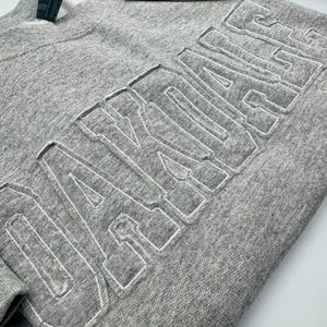 May include: A heather gray crewneck sweatshirt featuring the word OAKDALE in large, raised, block-style letters across the chest. The lettering is outlined with white stitching, creating a textured, dimensional effect on the fabric surface.