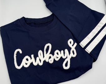May include: A navy blue long-sleeve sweatshirt featuring the word Cowboys in white textured chenille lettering across the chest. The sleeves are accented with two horizontal white stripes near the cuffs and a ribbed crew neckline.
