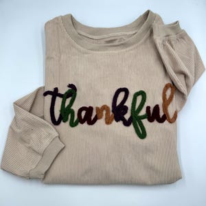 May include: A beige long-sleeve ribbed shirt featuring the word thankful embroidered in a cursive font across the chest. The letters are rendered in alternating colors of dark purple, forest green, and light brown textured yarn.