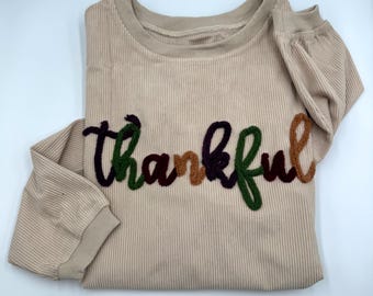 May include: A beige long-sleeve ribbed shirt featuring the word thankful embroidered in a cursive font across the chest. The letters are rendered in alternating colors of dark purple, forest green, and light brown textured yarn.
