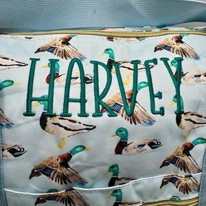 May include: Pale blue tote bag featuring a duck pattern and the name "HARVEY" embroidered in green. The bag has a light blue strap and a gold-coloured zip. The bag is ideal for carrying items.