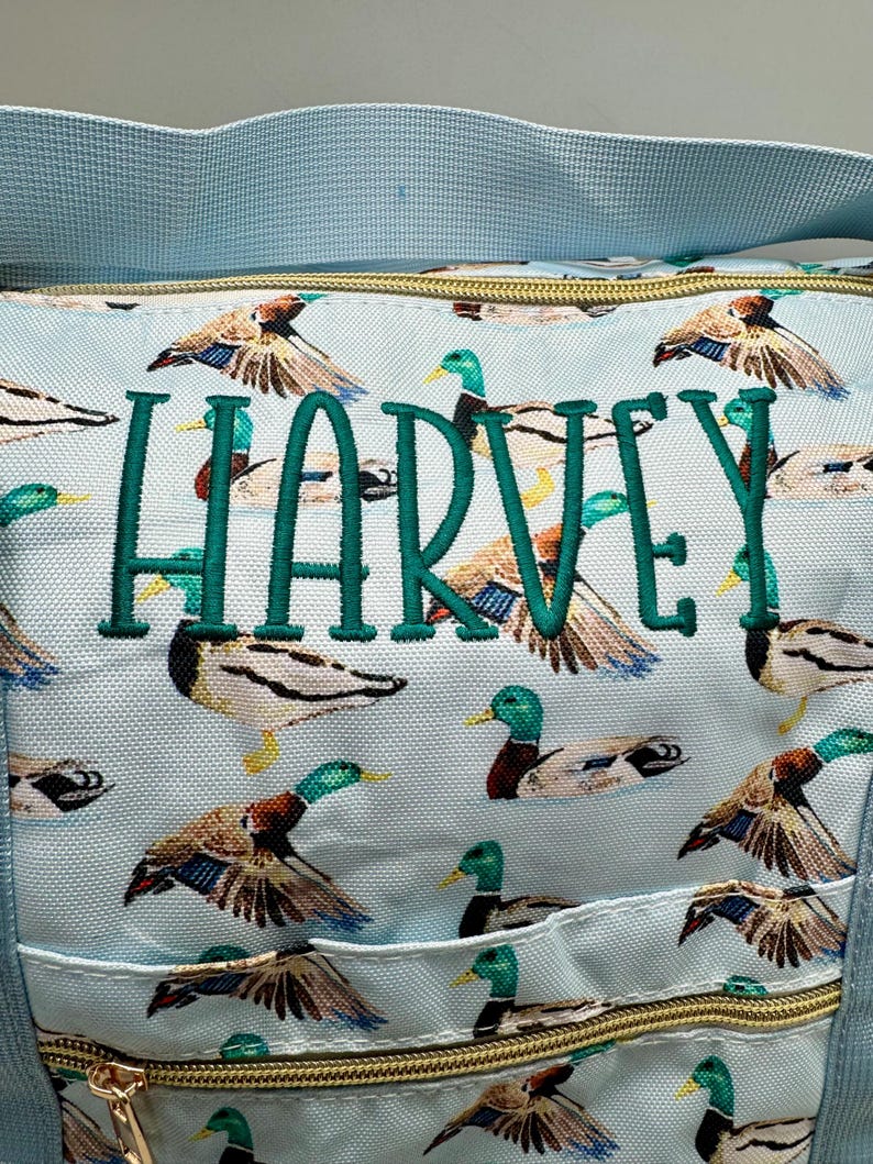 May include: Pale blue tote bag featuring a duck pattern and the name "HARVEY" embroidered in green. The bag has a light blue strap and a gold-coloured zip. The bag is ideal for carrying items.