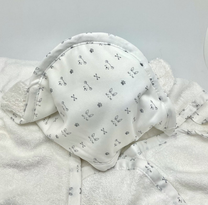 May include: White hooded baby towel with a pattern of gray bunny faces, paw prints, and crossbones. The towel is made of soft, absorbent material, perfect for after bath time. The hood is lined with the same patterned fabric.