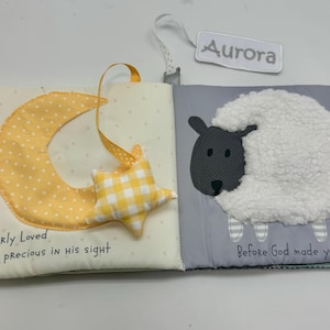 May include: Open cloth book with a yellow crescent moon and star, and a sheep illustration. The moon has white polka dots, and the star is yellow and white checkered. The text "Dearly Loved and precious in his sight" is on the left page. The right page has a sheep and the text "Before God made you".