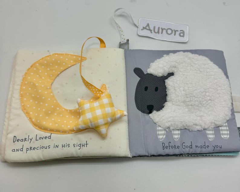 May include: Open cloth book with a yellow crescent moon and star, and a sheep illustration. The moon has white polka dots, and the star is yellow and white checkered. The text "Dearly Loved and precious in his sight" is on the left page. The right page has a sheep and the text "Before God made you".