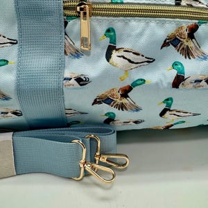 May include: A light blue duffel bag with a repeating pattern of mallard ducks. The bag features a gold zip, gold-coloured metal hardware, and a matching light blue shoulder strap.