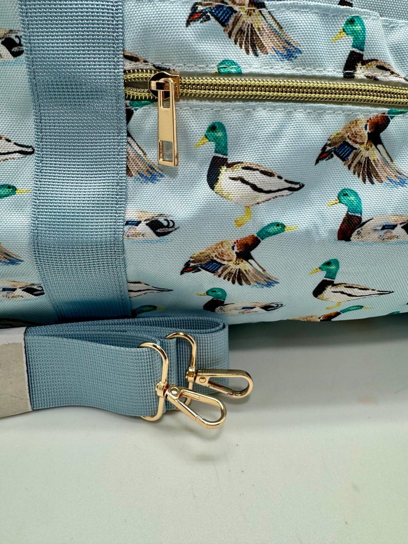 May include: A light blue duffel bag with a repeating pattern of mallard ducks. The bag features a gold zip, gold-coloured metal hardware, and a matching light blue shoulder strap.