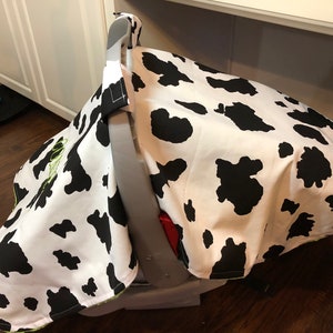 May include: A white and black cow print car seat cover with a green embroidered name. The cover is made of soft fabric and is designed to protect the car seat from dirt and spills.