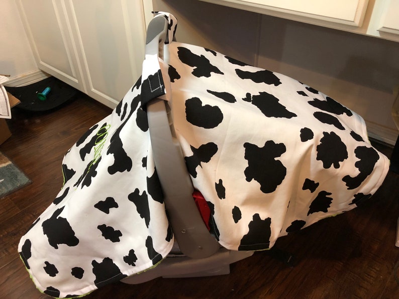May include: A white and black cow print car seat cover with a green embroidered name. The cover is made of soft fabric and is designed to protect the car seat from dirt and spills.