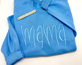 May include: A light blue sweatshirt with the word "mama" embroidered in a lighter shade of blue. A small heart is embroidered next to the word. The sweatshirt has long sleeves and a crew neck. The text "@CUSTOMTHREADSSHOP" is on a small wooden tag.