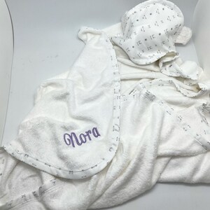 May include: White hooded baby towel and matching bonnet. The towel has a purple embroidered name "Nora" and a patterned border. The bonnet features the same pattern. The set is made of soft, absorbent material.