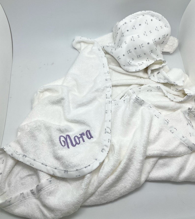 May include: White hooded baby towel and matching bonnet. The towel has a purple embroidered name "Nora" and a patterned border. The bonnet features the same pattern. The set is made of soft, absorbent material.
