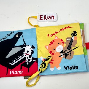 May include: A colorful cloth book for babies and toddlers with a piano, a violin, and a dog. The book has the words "Plinka Plink!" and "Squeak...squeak..." on the front and back covers. The book also has the words "Piano" and "Violin" on the pages.