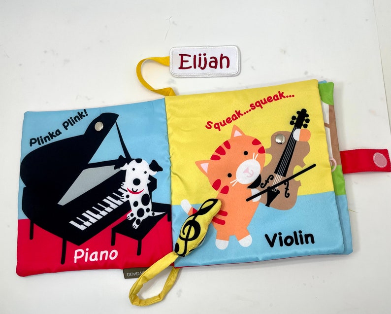 May include: A colorful cloth book for babies and toddlers with a piano, a violin, and a dog. The book has the words "Plinka Plink!" and "Squeak...squeak..." on the front and back covers. The book also has the words "Piano" and "Violin" on the pages.