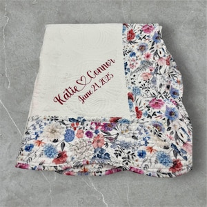 May include: Cream-colored quilted blanket with floral patterned border. The blanket features the embroidered names "Katie & Connor" with a heart symbol and the date "June 21, 2025" in burgundy thread. The floral pattern includes blue, pink, and white flowers.