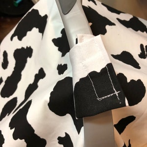 May include: A black and white cow print car seat cover with a gray handle. The cover has a white fabric strap with black stitching.