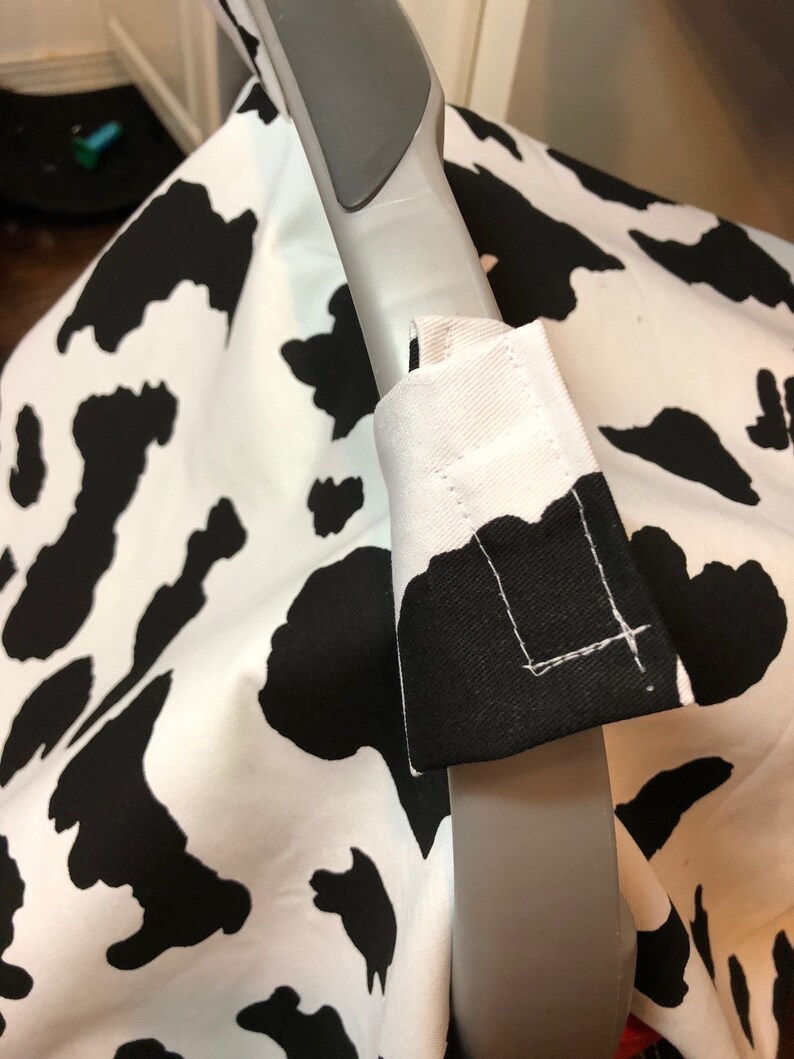 May include: A black and white cow print car seat cover with a gray handle. The cover has a white fabric strap with black stitching.