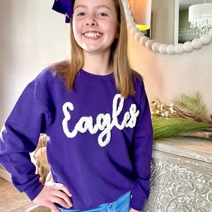 May include: A young person wearing a purple sweatshirt with the word "Eagles" in white fluffy letters. The sweatshirt has long sleeves and a crew neck. The person is wearing blue jeans and a purple bow in their hair.