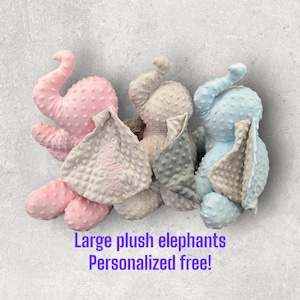 May include: Three large plush elephant toys, one pink, one gray, and one blue. Each elephant has a soft, textured fabric and a blanket attached. The text "Large plush elephants Personalized free!" is visible below the toys.