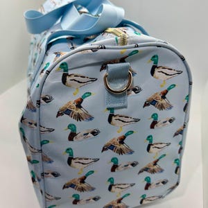 May include: A light blue duffel bag featuring a pattern of mallard ducks. The ducks are depicted with green heads, brown wings, and yellow feet. The bag has light blue handles and a gold-coloured zip.