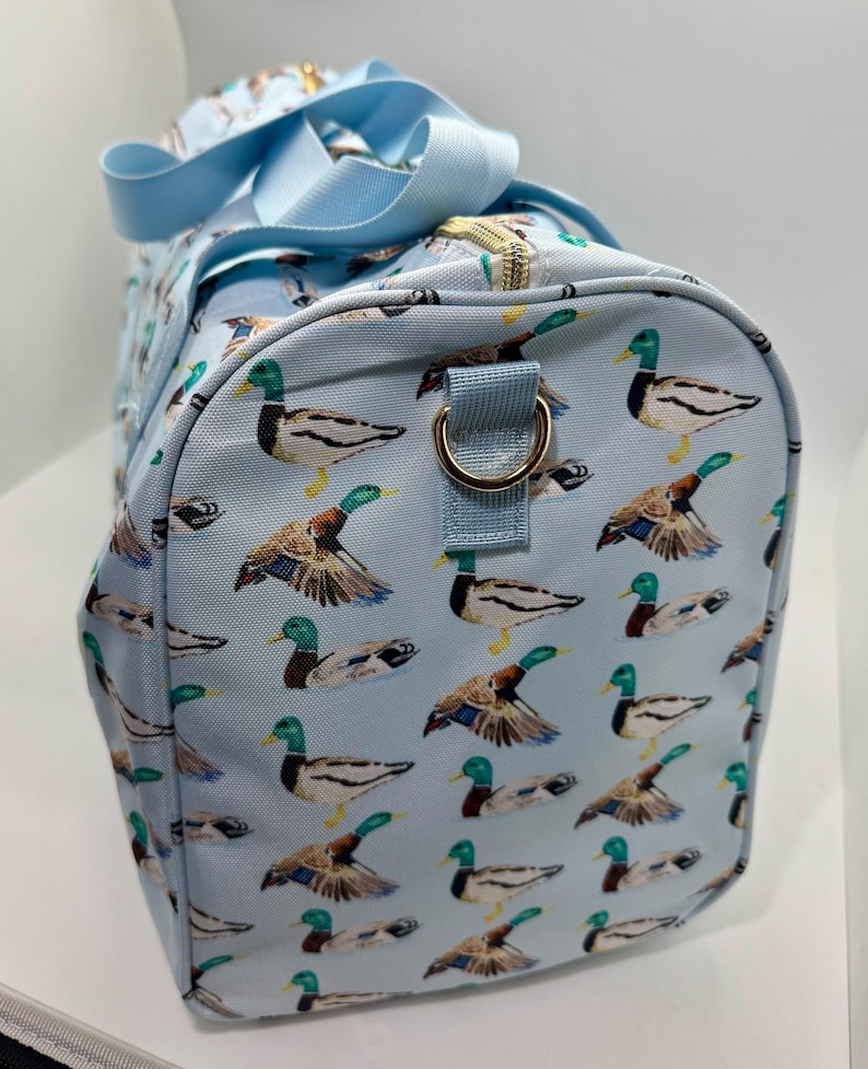 May include: A light blue duffel bag featuring a pattern of mallard ducks. The ducks are depicted with green heads, brown wings, and yellow feet. The bag has light blue handles and a gold-coloured zip.
