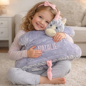 May include: A plush, lavender-colored cloud-shaped blanket with the name "NATALIE" embroidered in pink. The blanket has a fluffy border and a pink twisted tail. A stuffed unicorn head is attached to the blanket.