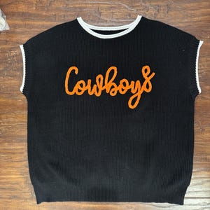 May include: A black sleeveless knit sweater vest featuring a crew neckline with white trim. The armholes also have white trim. The word Cowboys is embroidered in orange cursive script across the center of the chest on the front of the garment.