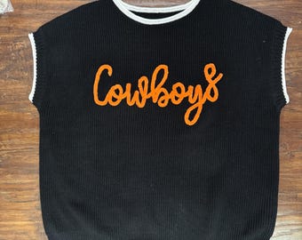 May include: A black sleeveless knit sweater vest featuring a crew neckline with white trim. The armholes also have white trim. The word Cowboys is embroidered in orange cursive script across the center of the chest on the front of the garment.