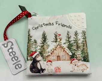 May include: A soft fabric book titled Christmas Friends featuring a winter forest scene with a bear, rabbit, and deer wearing Santa hats near a wooden cabin. A red ribbon with white polka dots attaches a white rectangular tag embroidered with Steele.
