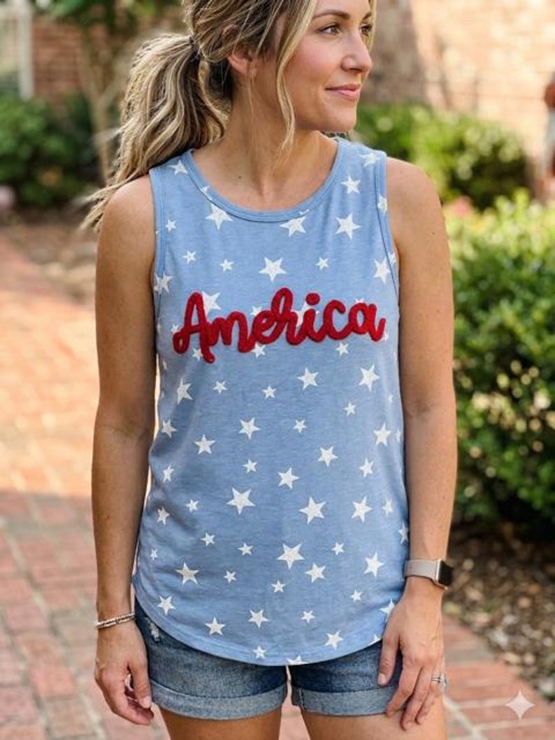 May include: A light blue sleeveless tank top featuring a repeating pattern of white stars and the word America written in red cursive script across the chest. The garment has a round neckline and a curved hemline, constructed from a soft fabric.