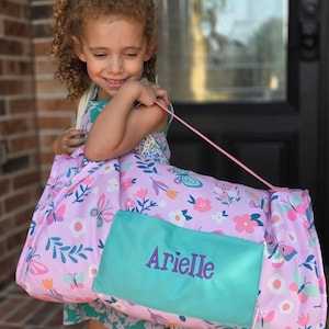 May include: A rolled-up, pink and purple mat with a butterfly and floral pattern, featuring a turquoise pocket embroidered with the name "Arielle" in purple. The mat has a pink strap for carrying.