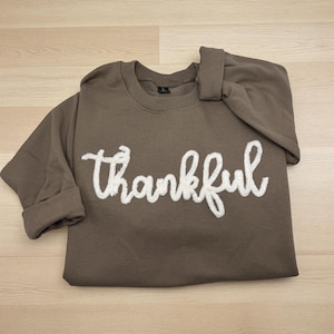 May include: A brown sweatshirt with the word "thankful" embroidered in white, cursive lettering. The sleeves are rolled up, and the sweatshirt is folded. The background is a light wood grain.