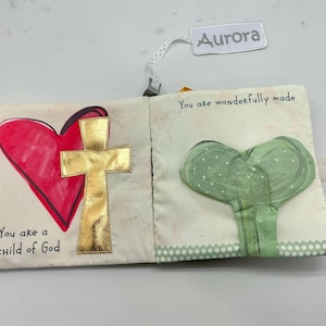 May include: Cloth book with a red heart and gold cross on one page, and a green heart-shaped design on the other. The text "You are a child of God" and "You are wonderfully made" are printed on the pages. A tag that says "Aurora" is attached.