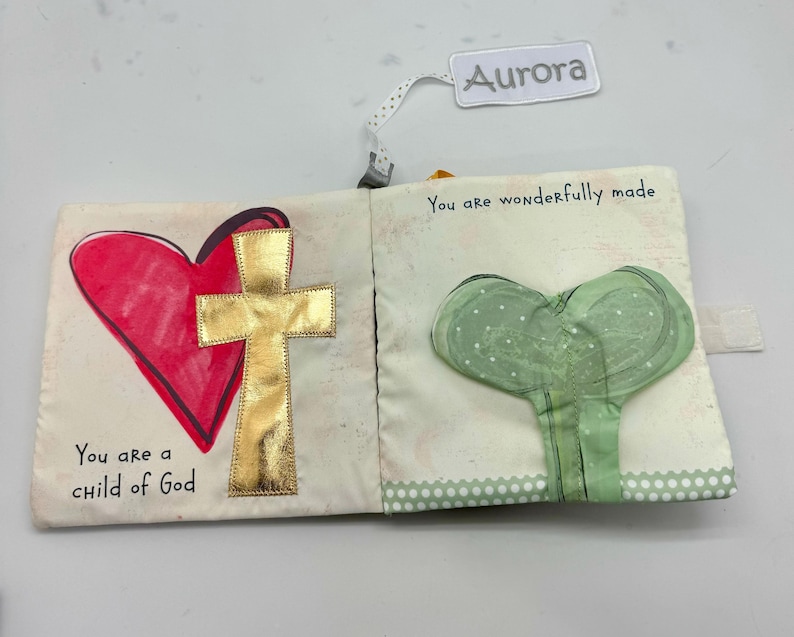 May include: Cloth book with a red heart and gold cross on one page, and a green heart-shaped design on the other. The text "You are a child of God" and "You are wonderfully made" are printed on the pages. A tag that says "Aurora" is attached.
