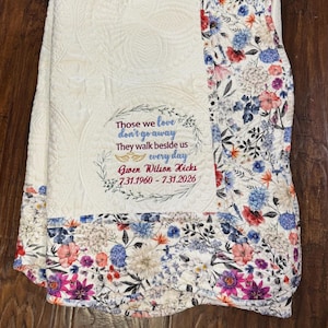 May include: A cream-colored quilted throw blanket featuring a floral border with blue, pink, and purple flowers. The center panel includes embroidered text: Those we love don't go away They walk beside us every day Gwen Wilson Hicks 7.31.1960 - 7.31.2026.