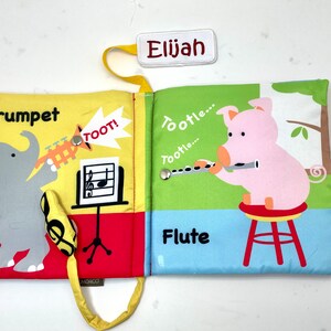 May include: A yellow, red, and blue fabric book with a pig playing a flute and an elephant playing a trumpet. The book has a yellow strap and a tag that says "Elijah".