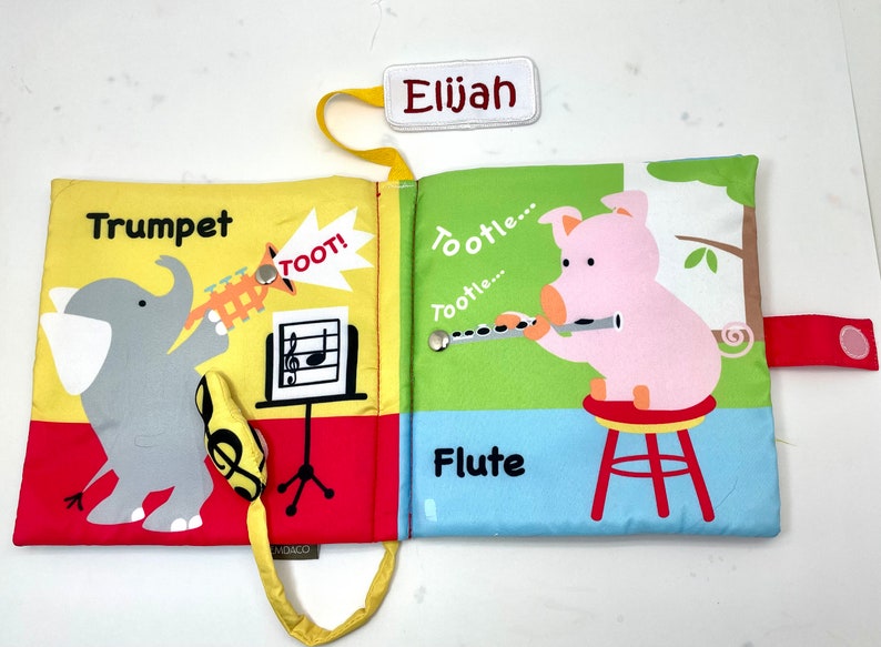 May include: A yellow, red, and blue fabric book with a pig playing a flute and an elephant playing a trumpet. The book has a yellow strap and a tag that says "Elijah".