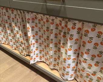 May include: A white fabric valance with a repeating pattern of orange floral designs and green leaves. The valance is draped under a grey cabinet with a gold-toned handle. The fabric has a scalloped edge.