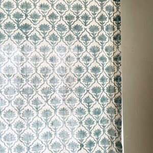 May include: A curtain panel featuring a repeating teal floral pattern on a white background. The fabric displays a symmetrical botanical motif arranged in a lattice design, with a dark green trim along the top edge and a metal mounting bracket.
