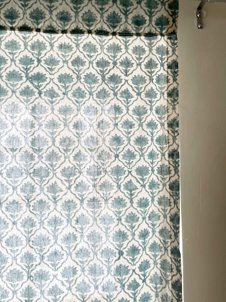 May include: A curtain panel featuring a repeating teal floral pattern on a white background. The fabric displays a symmetrical botanical motif arranged in a lattice design, with a dark green trim along the top edge and a metal mounting bracket.