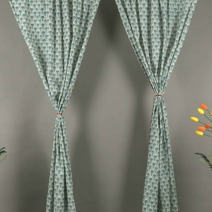 May include: A pair of floor-length curtains featuring a repeating teal and white floral pattern. The fabric is gathered at the center of each panel with a thin red tie, creating a draped effect that hangs from a rod at the top of the panels.